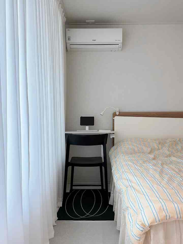 Minimal white-toned small bedroom featuring sheer curtains on left, a compact desk with devices center-left, and bed on right side