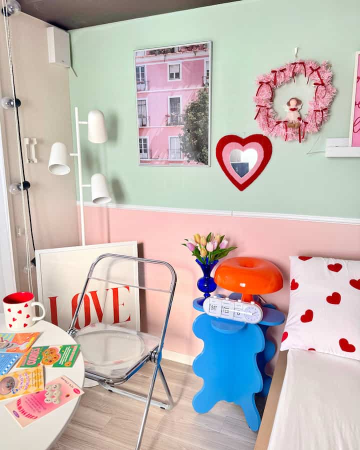 ミント色とピンク色の壁がある小さな一人暮らし空間、透明な椅子と青いサイドテーブルに置かれた時計と花瓶が配置された可愛らしいインテリア