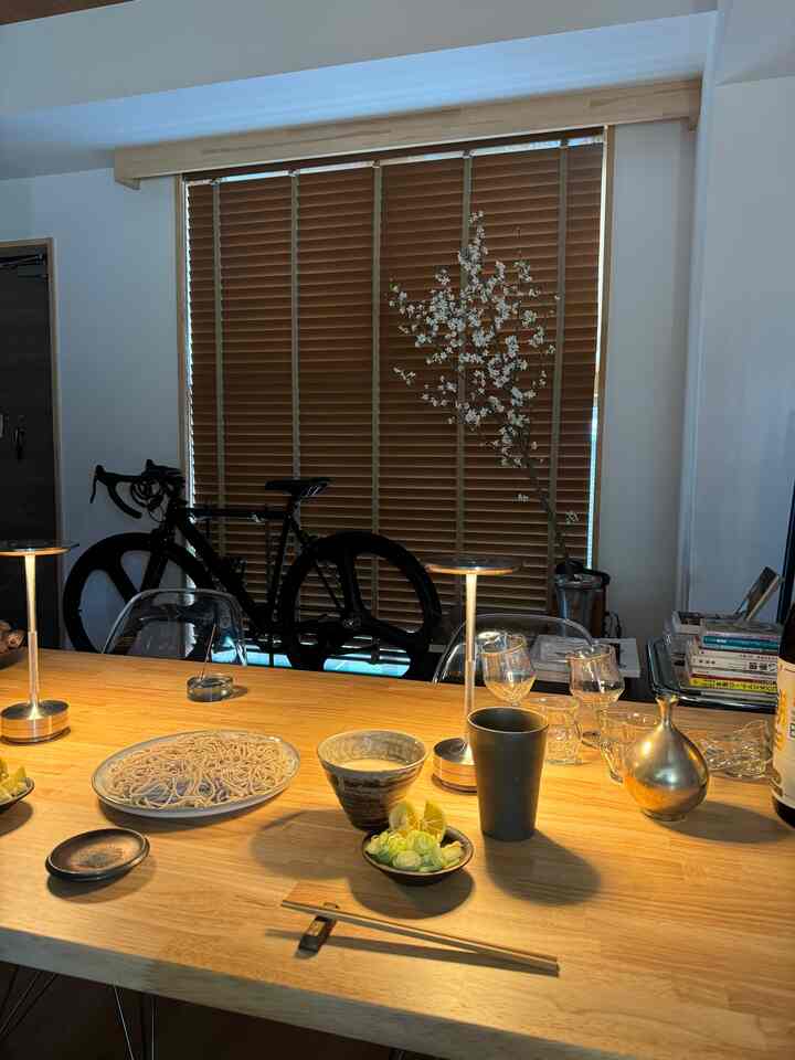 ブラウンの木目調ブラインドがあるコンパクトな一人暮らし1LDK空間で、室内自転車や食事の準備がされたテーブルが配された雰囲気のあるインテリア