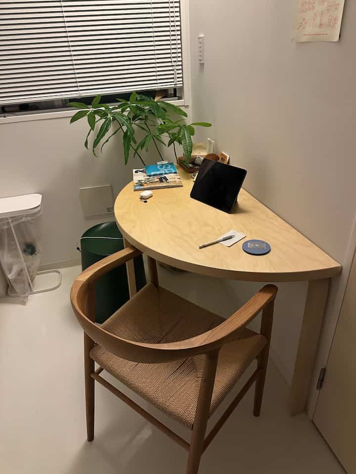 Natural tone small single household room featuring a corner wooden desk, armchair, and green plant in a clean setup
