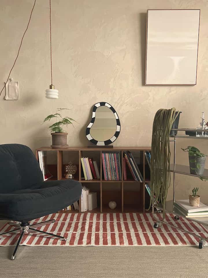 ベージュの壁と赤いストライプのラグがあるリビング、黒いアームチェアと木製の本棚が配置された自然な雰囲気のインテリア