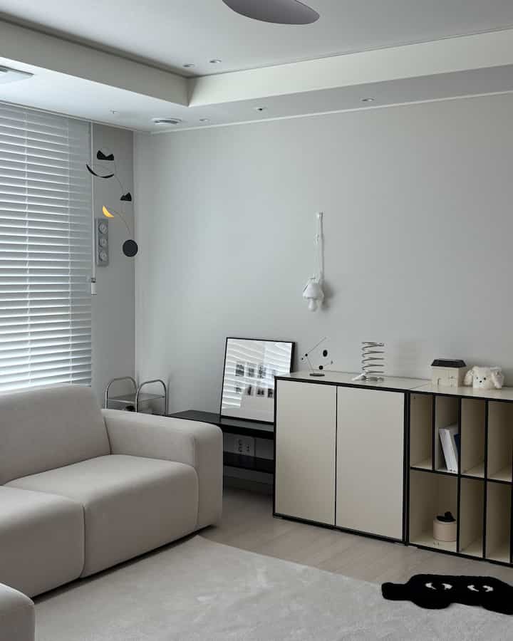 A white and beige toned living room featuring a simple sofa, hanging mobile, and sleek storage cabinet with a minimal modern atmosphere