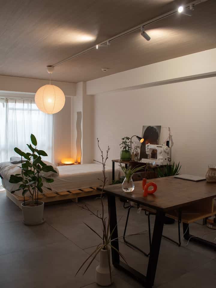 温かみのある木目調と白を基調としたワンルーム寝室、ベッドとデスク、植物が配置された居心地の良いジャパンディスタイルのインテリア