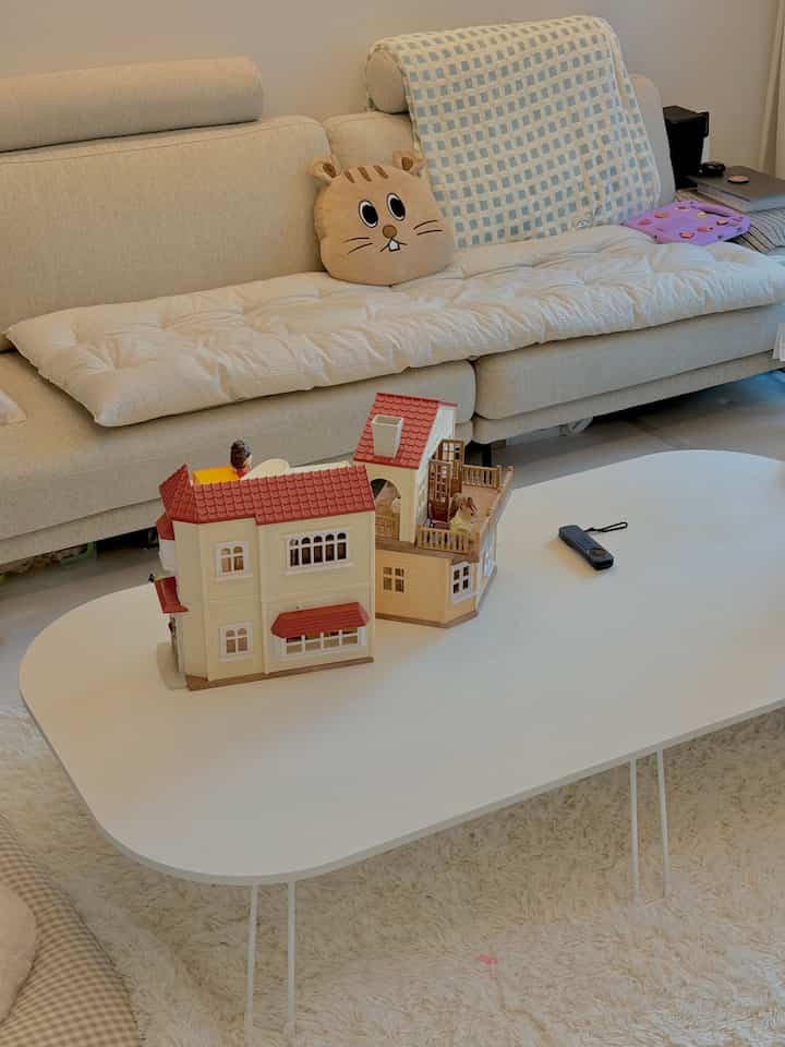 ベージュとホワイトの色調のリビング、中央にホワイトのコーヒーテーブルと子供のおもちゃの家、ベージュのソファが配置された居心地の良い空間