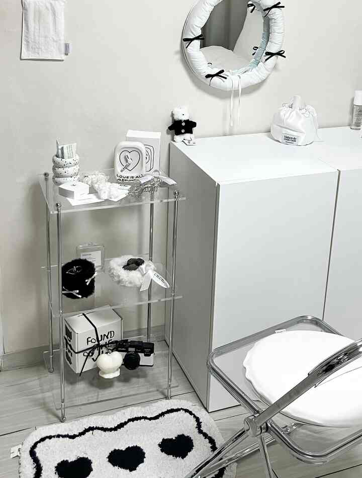 A clean monotone room with white walls, mid-century modern acrylic shelving, a round mirror decorated with ribbons, and a black and white heart rug
