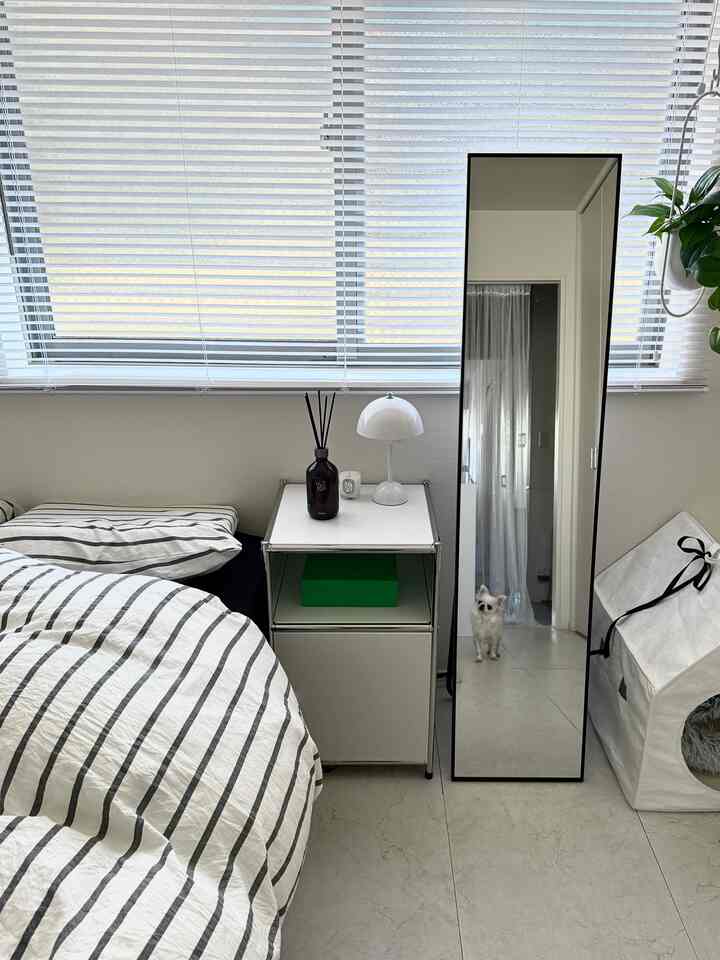 モノトーン調のシンプルな寝室、ストライプ柄の寝具と白いナイトテーブル、全身鏡が配置された雰囲気のあるインテリア