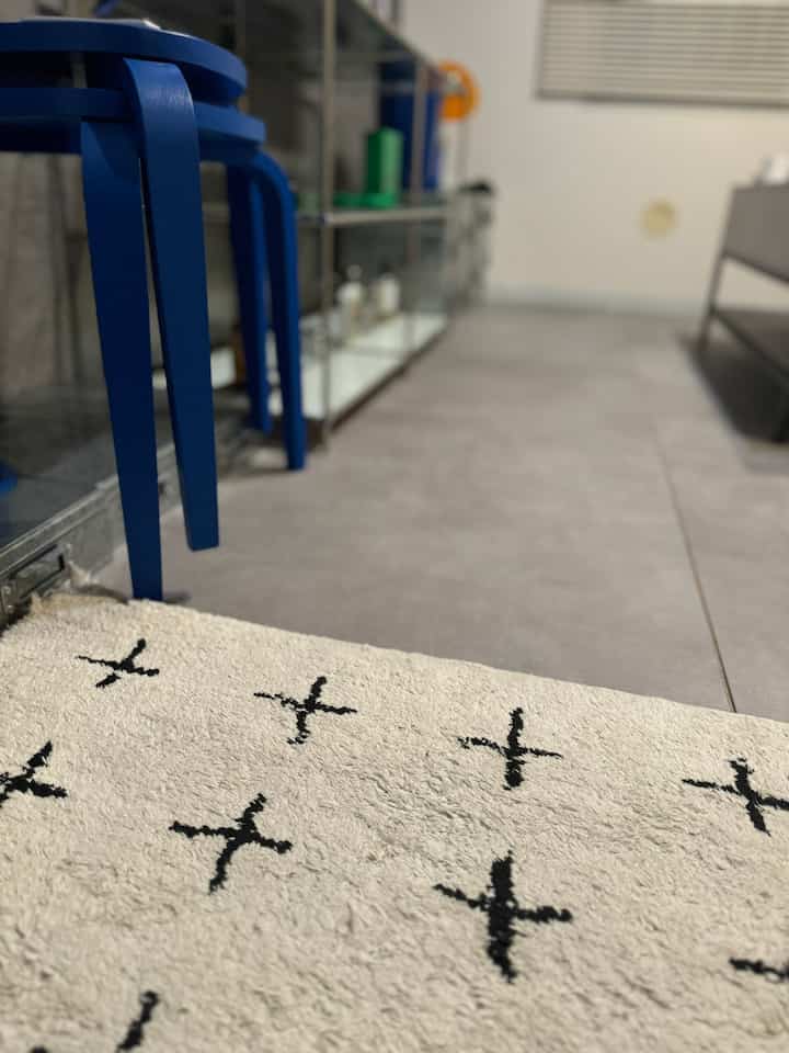 グレーのフロアタイルの床に青いスツールと黒い十字柄の白いラグが配置されたシンプルな玄関空間