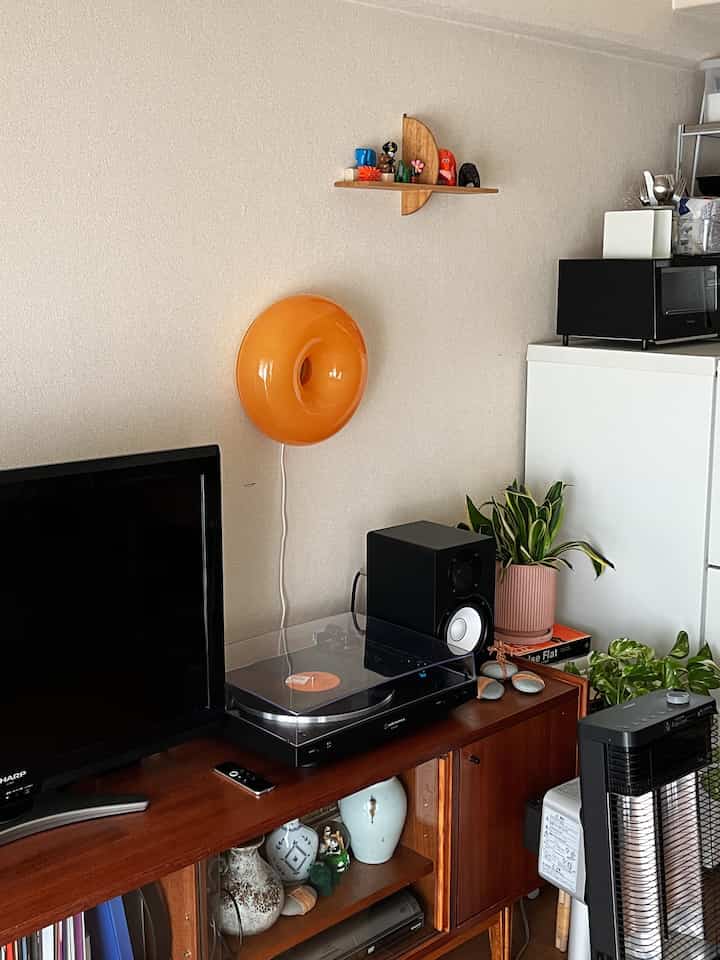 アイボリーの壁とブラウンのヴィンテージなテレビ台があるリビング、オレンジの円形ウォールランプ、ターンテーブル、植物が配置された雰囲気のある空間