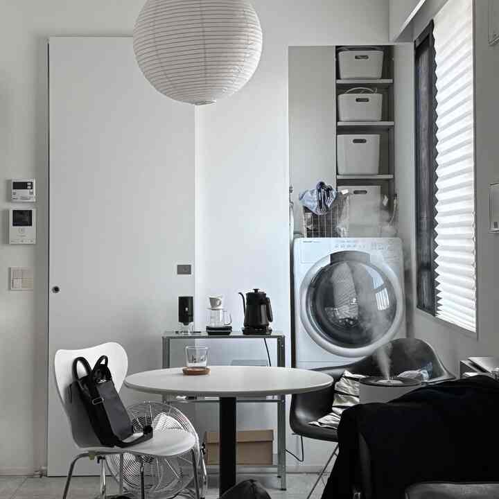 モノトーンの狭く縦長の部屋、丸いダイニングテーブルと椅子、洗濯機とコーヒー用品が配置された実用的なインテリア