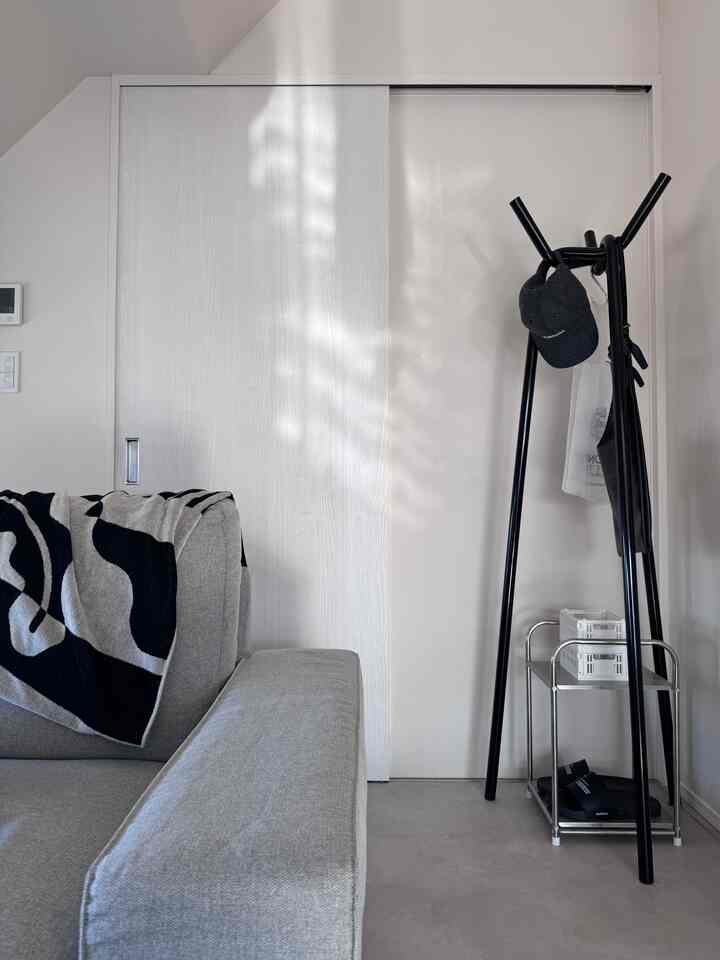 白い壁と淡い床の調和が取れたリビング、グレーのソファと黒いコートラックが配置されたミニマルな空間