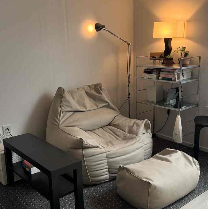 ベージュのビーンバッグソファとグレーのラグ、メタルシェルフとシンプルな照明が配置されたミニマルなリビングルーム