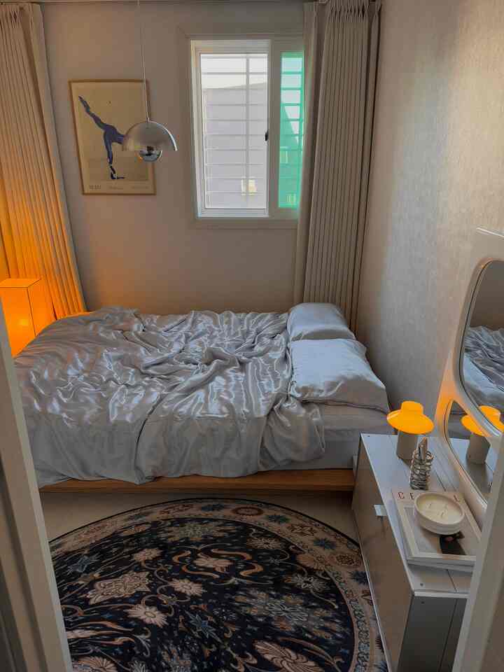 スカイブルーの色調の寝室、中央に白いサテンの掛け布団カバーをかけたベッドとベージュのカーテン、円形のペルシャ絨毯が置かれた雰囲気のある空間