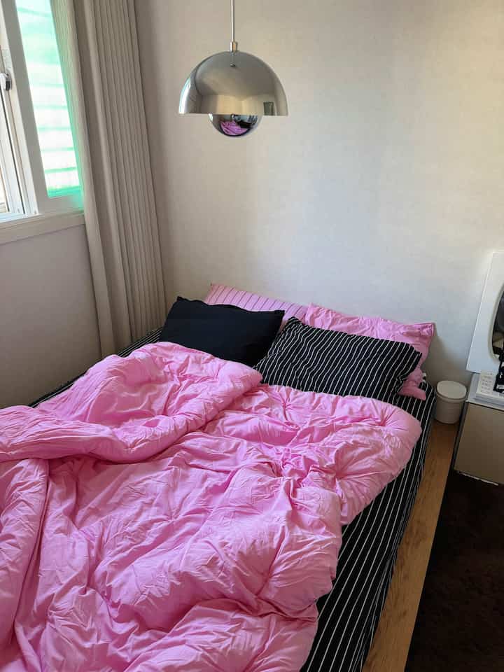 ピンクの寝具が印象的なモダンでナチュラルな寝室、木製ベッドとメタリックなペンダントライトが配置された雰囲気のあるインテリア