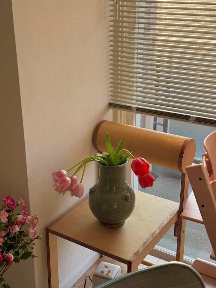 ナチュラルな木目調とベージュの壁、ブラインドが調和する狭いベランダの空間に、花瓶と花が置かれたシンプルなスツールが配置された落ち着いたコーナー