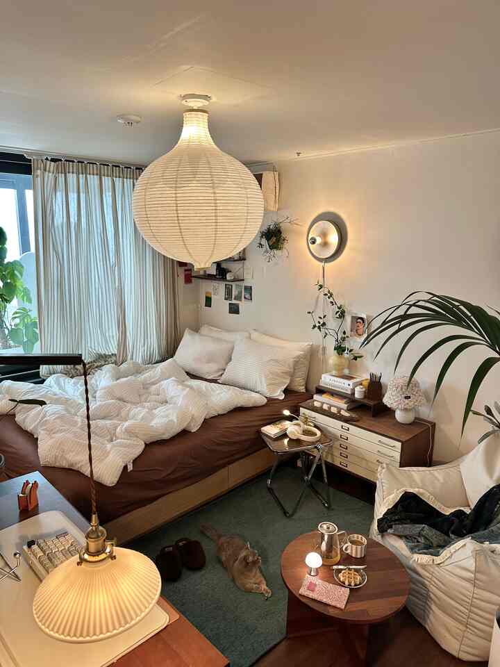 ブラウンとナチュラルな色調の寝室、木製ベッドと白い寝具、緑のラグ、多様な植物が配置された居心地の良い空間