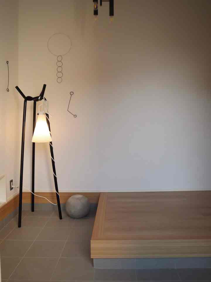 ベージュの壁と木目調のプラットフォームが調和したシンプルでモダンな玄関、ブラックのコートハンガーとホワイトのフロアランプがアクセントになっている