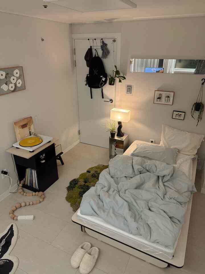 アイボリーとベージュの色調のワンルーム寝室、ミッドセンチュリーモダンのナイトテーブルと温かみのある照明が配置された落ち着いた空間