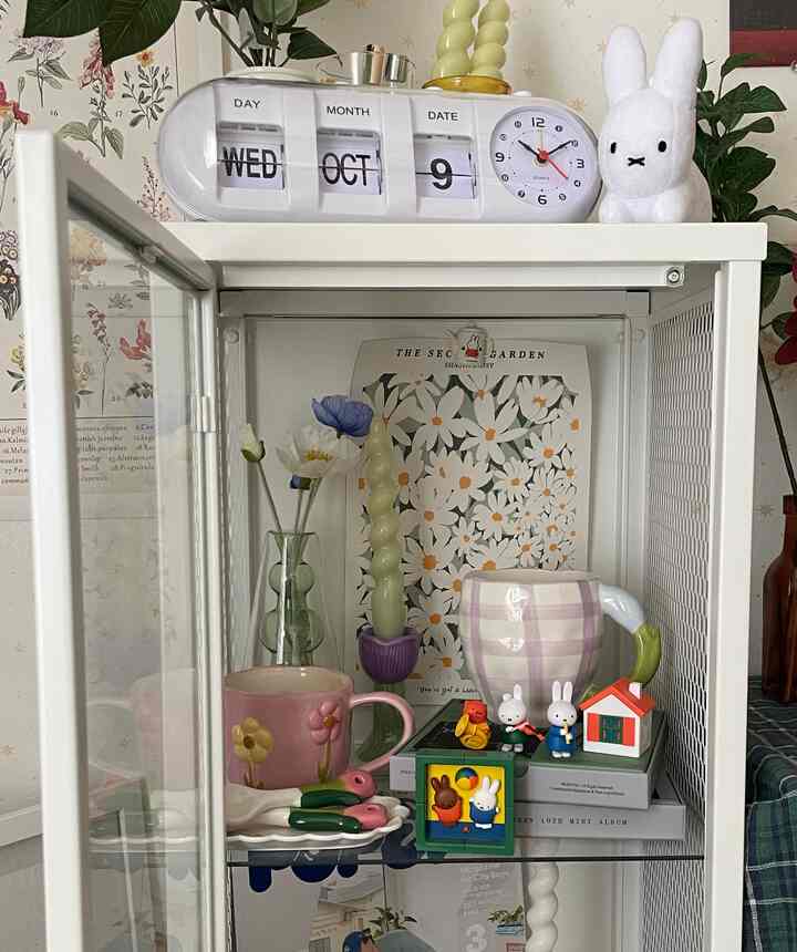 ホワイトフレームのガラスシェルフに花柄ポスター、チェック柄のマグカップ、キャンドルなどが配置されたかわいらしい韓国インテリアのディスプレイスペース