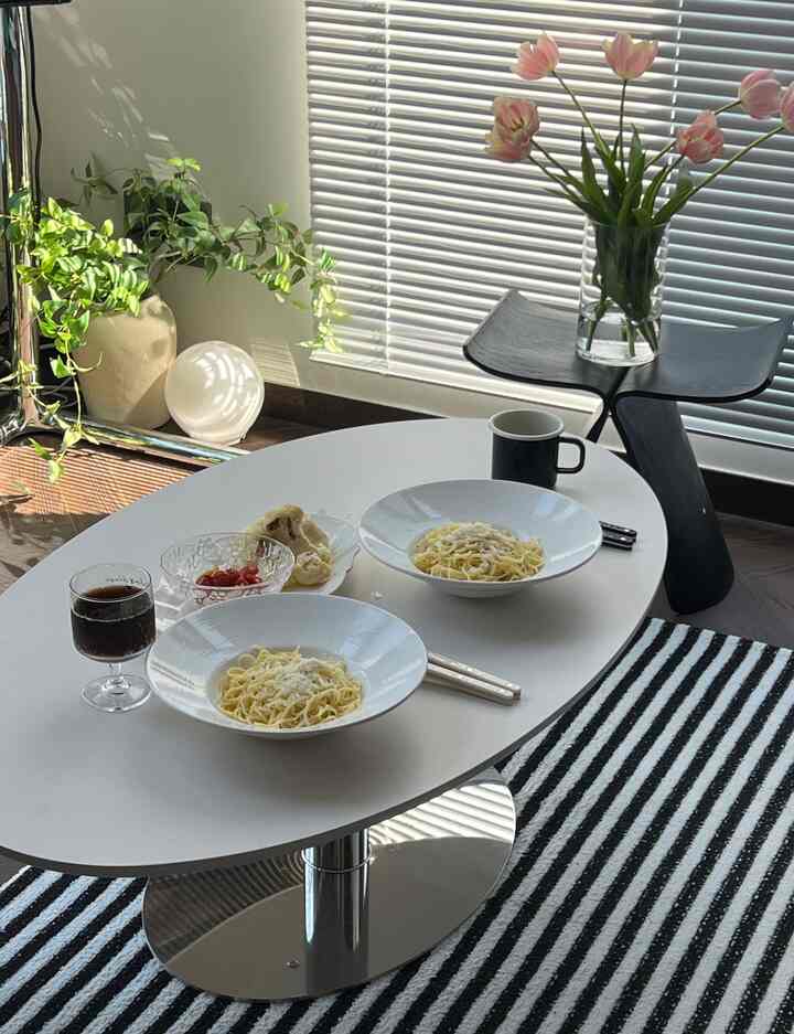 Modern white and black toned living room featuring oval coffee table with pasta dishes and decorative plants, creating a clean and natural atmosphere