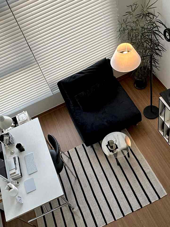 Natural wood floor and white blinds in a compact home office space featuring a black armchair and floor lamp with a modern atmosphere