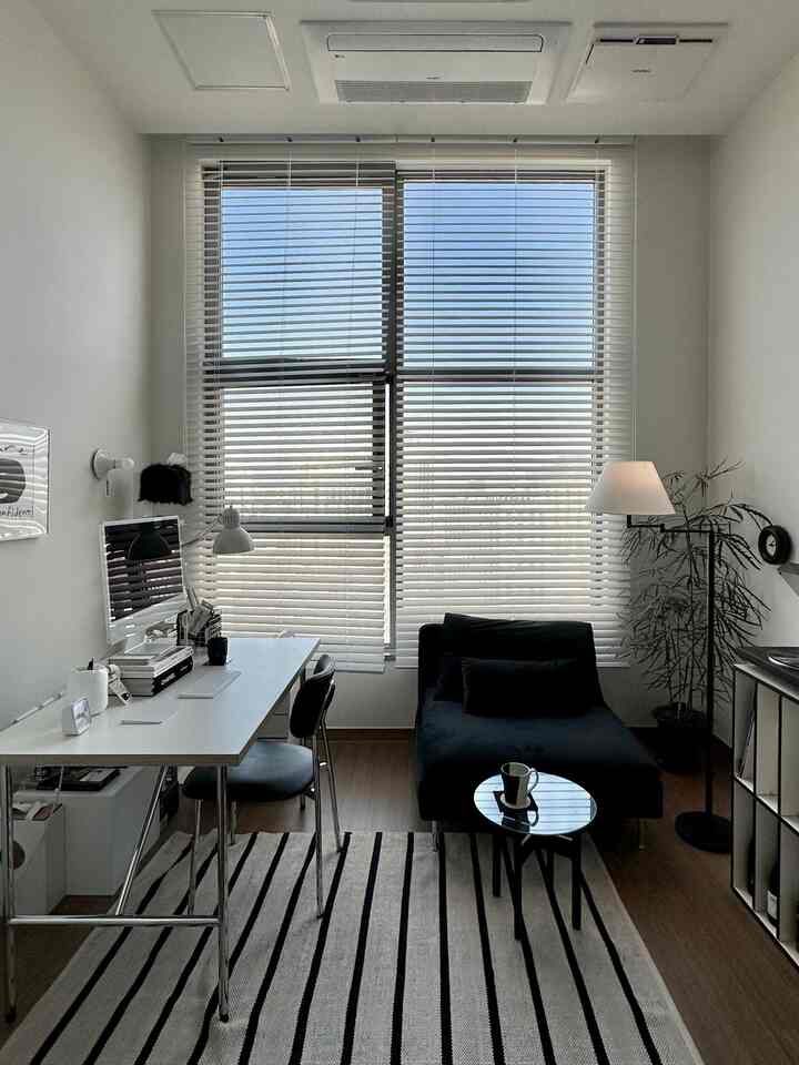 Modern home office in a narrow room with white walls and black furniture, featuring a desk, office chair, and window blinds