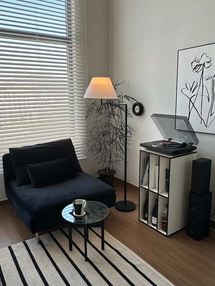 A stylish studio living room in white and black tones featuring a black single sofa, floor lamp, and turntable