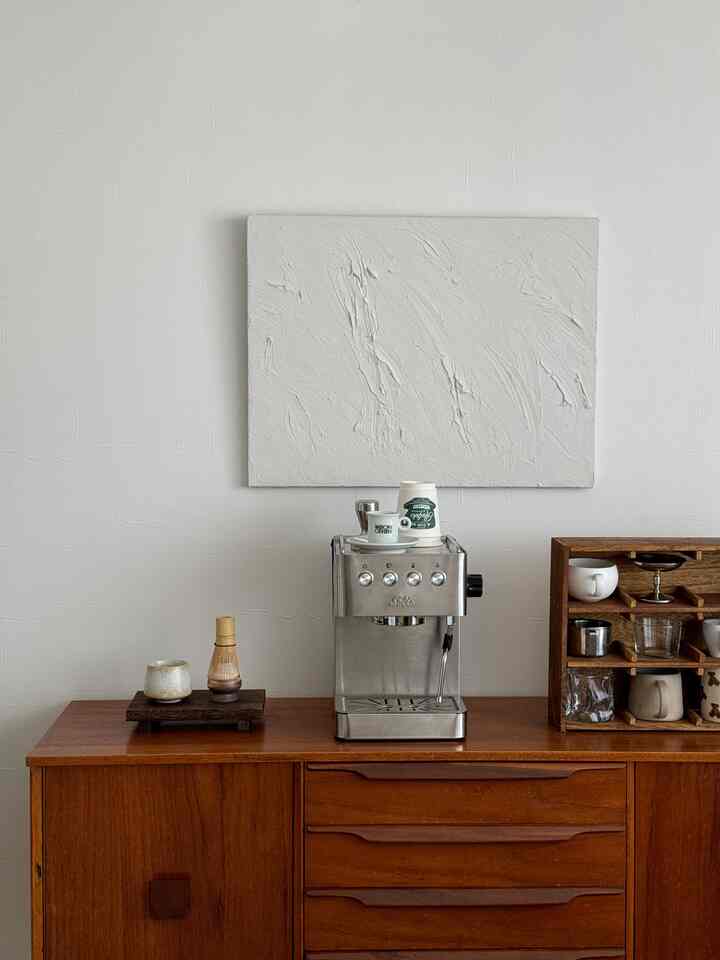 ナチュラルな木目調のサイドボードとステンレス製エスプレッソマシンが配置された白い壁の清潔感あるおうちカフェ