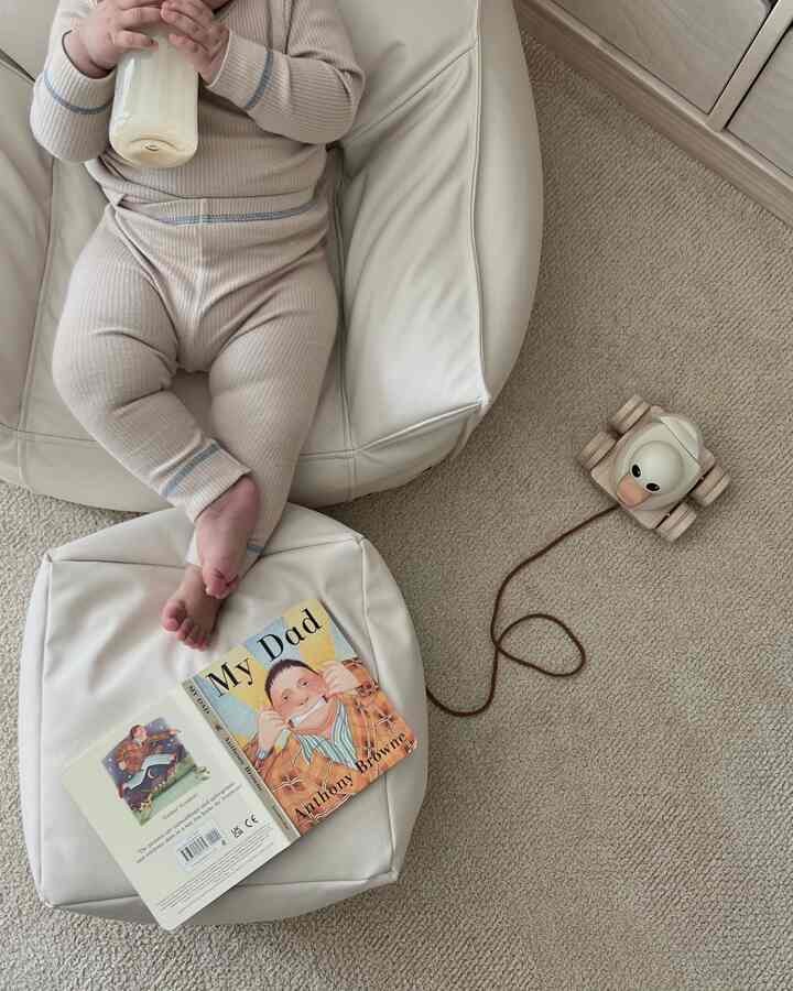 ベージュトーンの子供部屋の読書スペースで、白いビーズクッションに座る赤ちゃんがミルクを飲んでおり、木製のおもちゃとラグが配置された温かみのある空間
