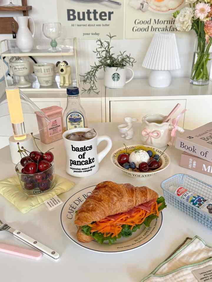 A white and pink toned home cafe space featuring a croissant sandwich and yogurt bowl on a dining table with a cozy atmosphere.