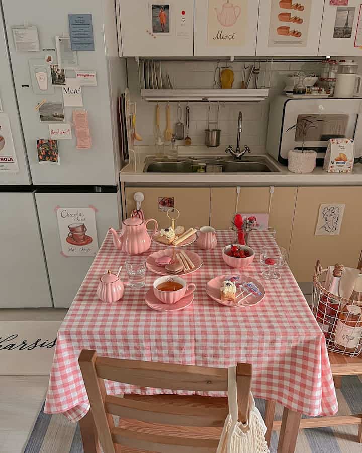 A cozy kitchen space in pink and beige tones featuring a checkered tablecloth with tea sets and snacks, evoking a home cafe vibe