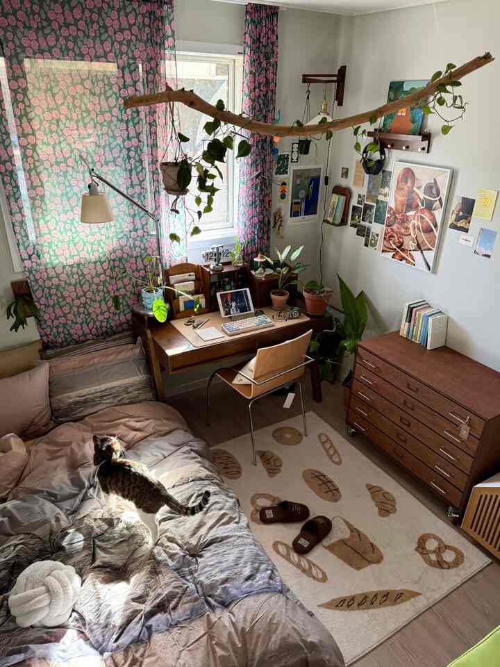 ナチュラルな色調のワンルーム寝室兼ホームオフィス、花柄カーテンと多くの植物が配置された温かみのあるインテリア