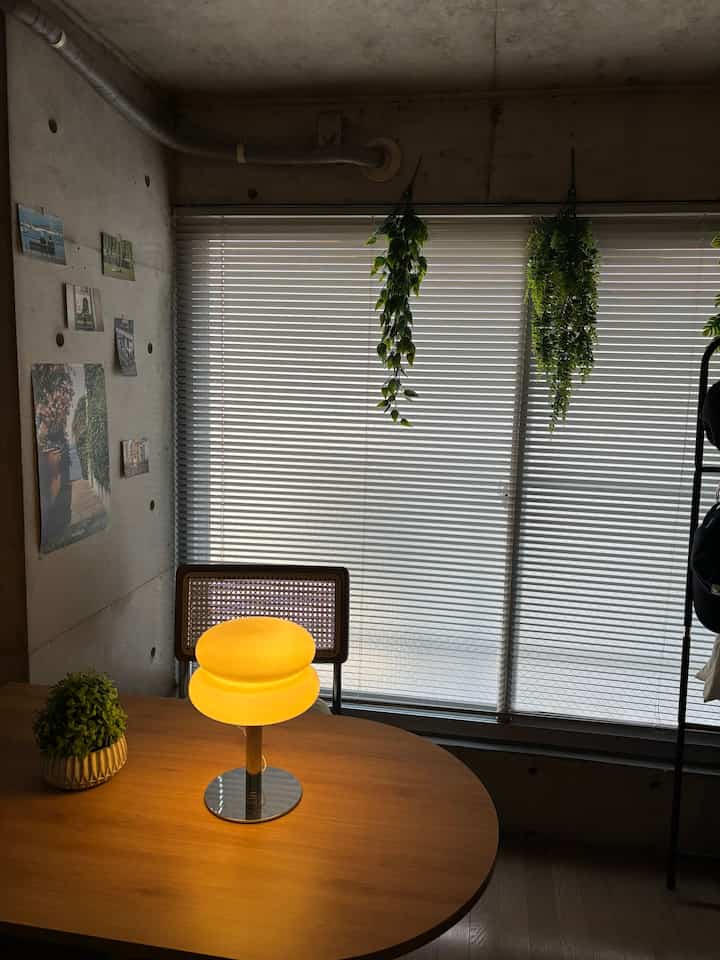 ナチュラルブラウンのダイニングテーブルに黄色いテーブルランプ、窓にブラインドと緑の植物が配置されたホテルライクなリビング空間