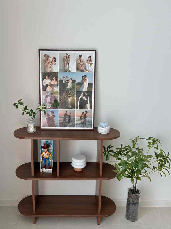 Brown wood-tone three-tier shelf with plants and framed photos against white walls and floor, creating a simple and natural bedroom corner