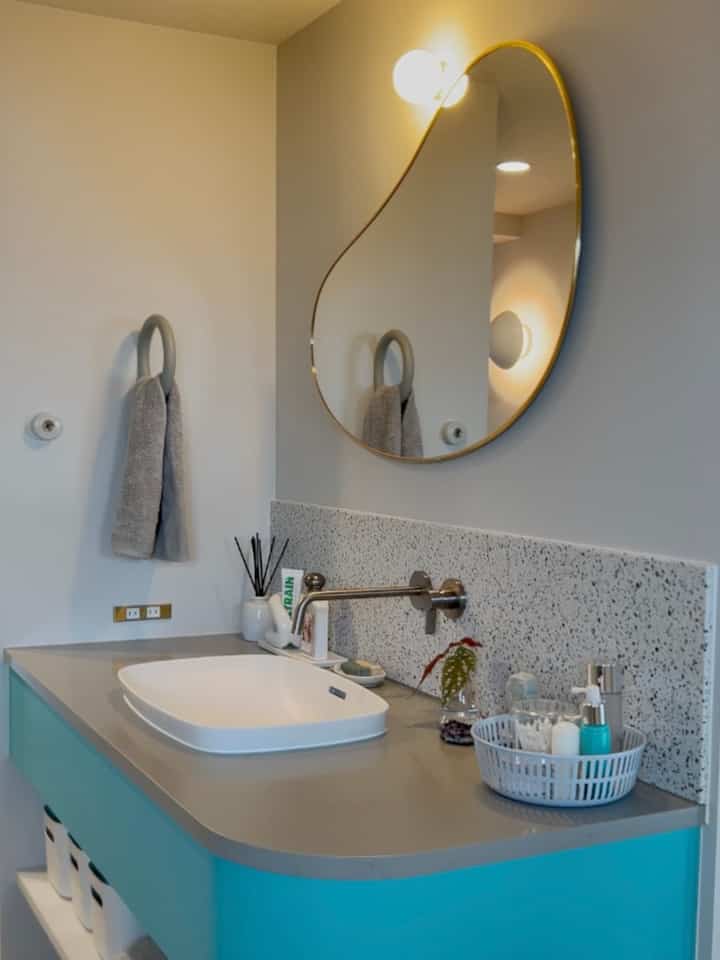 パステルブルーのトーンが特徴のバスルーム、独特な形のゴールドフレームミラーとスタイリッシュな洗面台が配置された清潔感ある空間