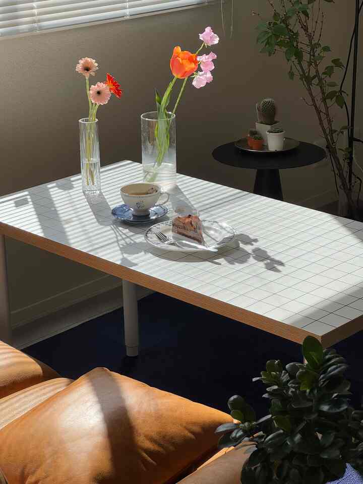 Modern living room with brown sofa and white tiled table, featuring vases of flowers and a home cafe setup with tea and cake
