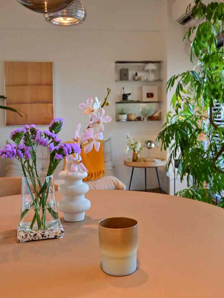 ナチュラルな色調のリビング、中央のテーブルに花飾りとカップが置かれ、周囲に植物が配置された雰囲気のある空間