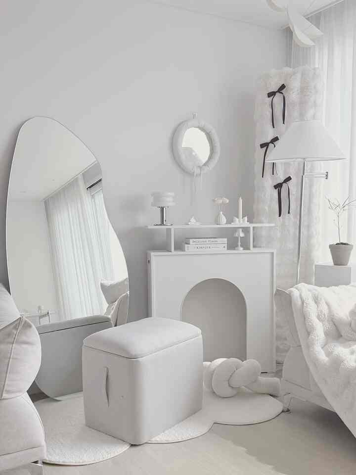 White-toned living room featuring a storage fabric stool at center, an irregular shaped mirror, and a floor lamp with a modern aesthetic