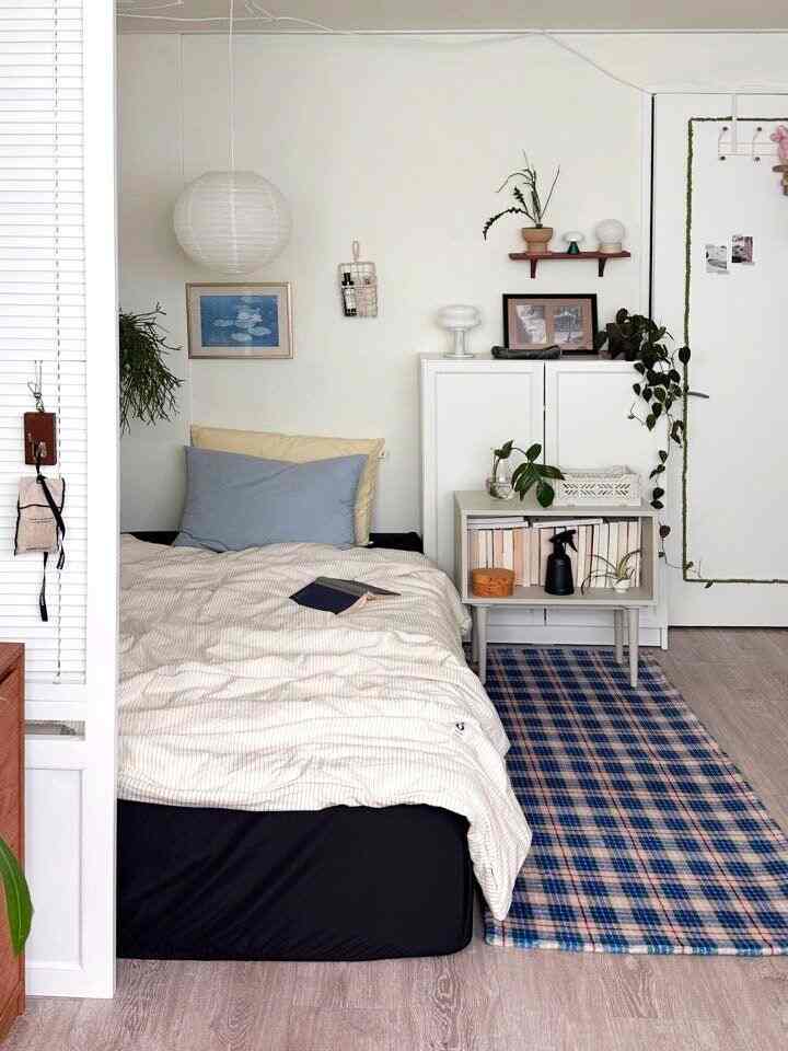 ナチュラルトーンの白い壁のワンルーム寝室、ベッドと収納棚、チェック柄のラグが配置された雰囲気のある空間