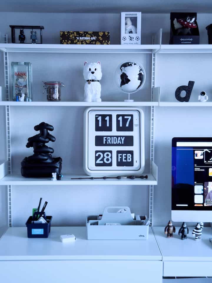 ホワイトとグレーのトーンのホームオフィス、時計や様々なオブジェが棚とデスクに配置された印象的な空間