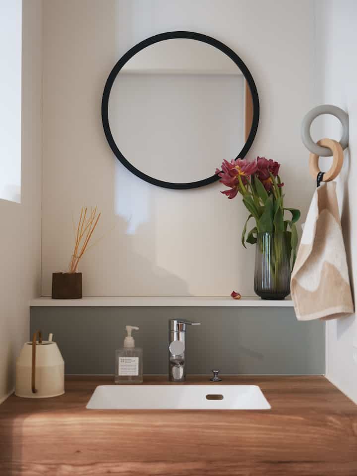 ナチュラルカラーの縦長洗面スペース、丸い黒い鏡と木目調の洗面台が調和したシンプルな印象のインテリア