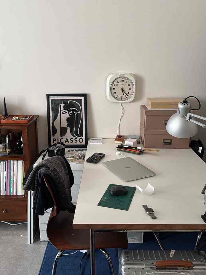 A white-walled, wood-tone mid-century modern home office featuring a clean desk, wall clock, and task lighting