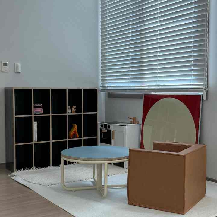 白と茶色のトーンの子供部屋、子供用ソファと3段の本棚、丸テーブル、キッチンおもちゃが配置された整頓された空間