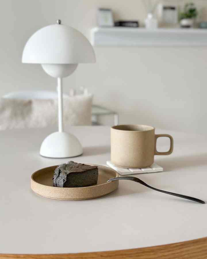 ナチュラルカラーのシンプルなおうちカフェ空間、ベージュのマグカップとプレートに乗った黒ゴマチーズケーキが置かれたテーブル