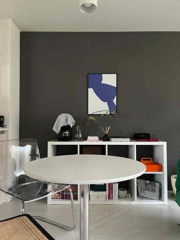 ホワイトとダークブラウンの壁が調和するダイニング、透明の椅子と丸テーブル、白い収納棚に青と白のポスターが配置されたモダンなインテリア