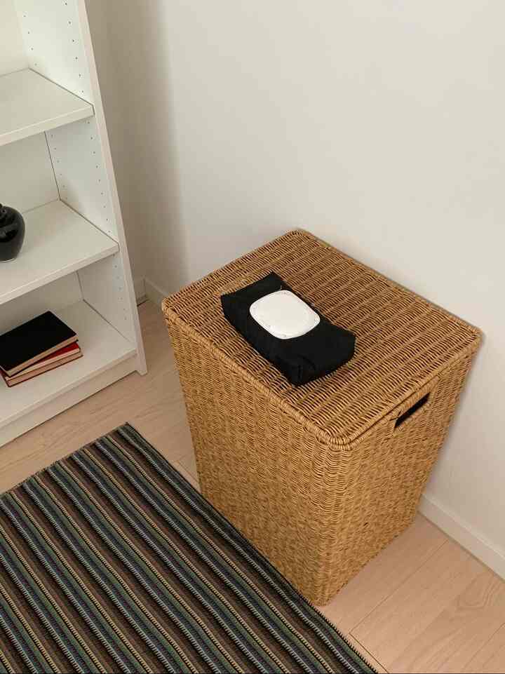 白い壁と本棚を背景に、ラタンの収納バスケットと黒いおしりふきカバーが置かれた自然でシンプルなインテリア
