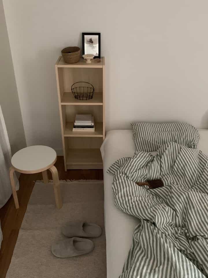 ナチュラルな色調の寝室、木目調の本棚とスツール、白い掛け布団カバーが配置されたシンプルなインテリア