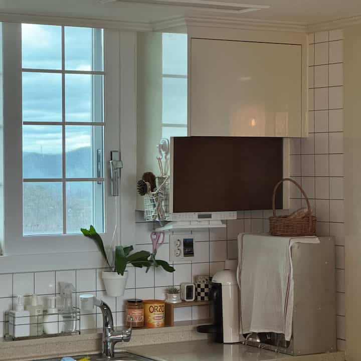 白とナチュラルトーンのキッチン、窓と流し台周辺にコーヒーマシンと収納バスケットが配された雰囲気のある空間