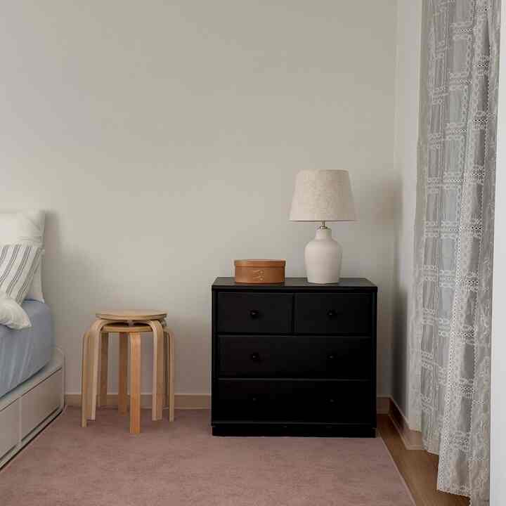 白い壁の寝室、黒いチェストにテーブルランプと木製スツールが置かれた、温かみのあるナチュラルな雰囲気のインテリア