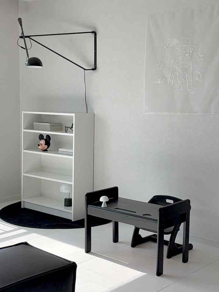 モノトーンのリビングコーナー、子供用机と白い本棚、壁照明が配置されたミニマルな印象の空間