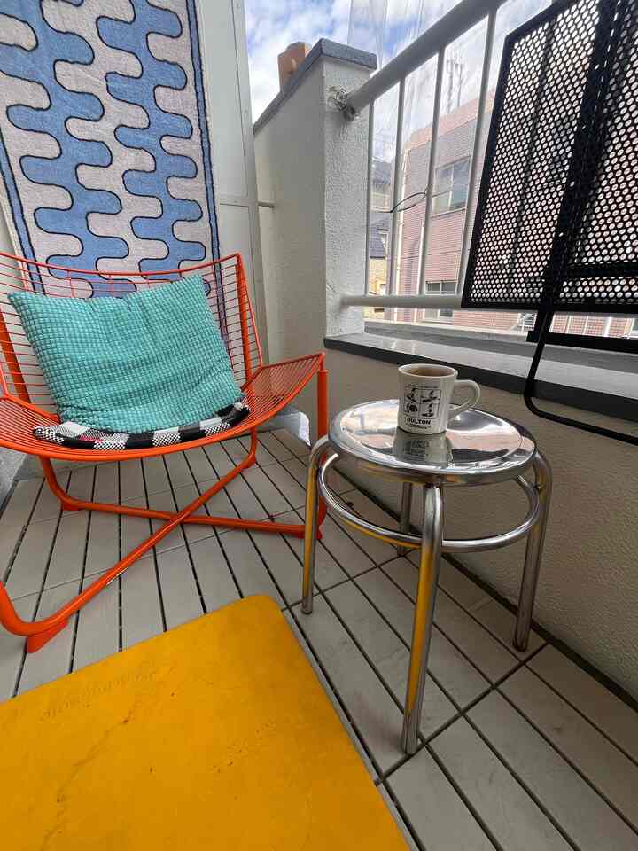 Gray floor and white balcony walls with an orange metal chair and silver stool in a simple modern studio apartment balcony space